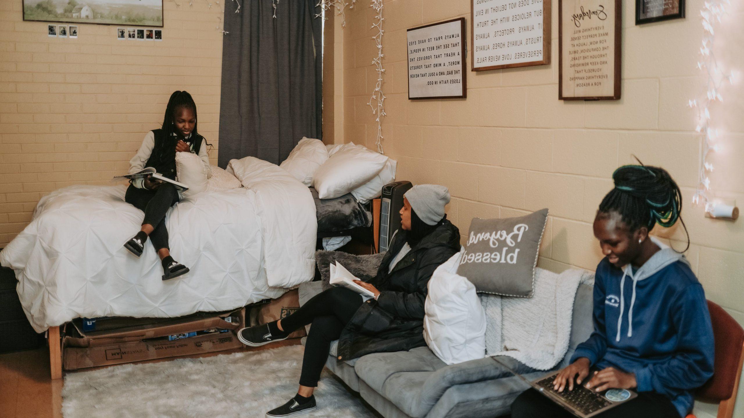 Three students in a dorm room; one is sitting on a bed, one is on a couch, one is in a chair and working on her laptop.